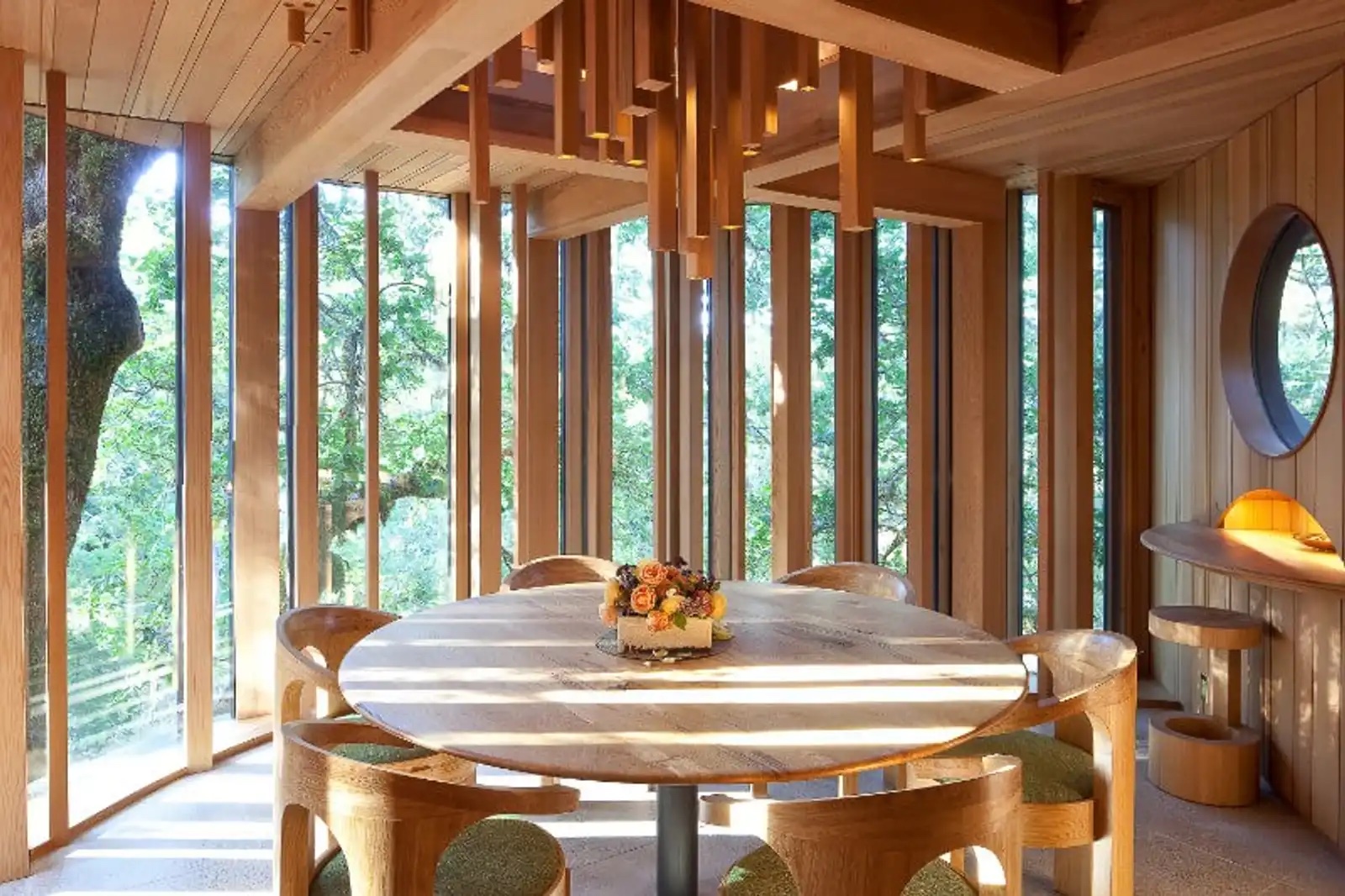 Round dining table beneath a beam-bundle pendant light, with tall slatted windows opening to the forest.